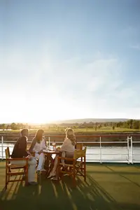Zwei Paare genießen ein Abendessen mit Wein auf dem Sonnendeck eines Flussschiffs bei Sonnenuntergang, umgeben von grüner Landschaft.