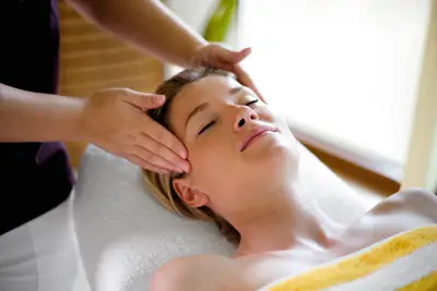 Eine entspannte Frau liegt auf einer Massageliege und erhält eine Gesichtsmassage von den Händen einer anderen Person in einem Spa.