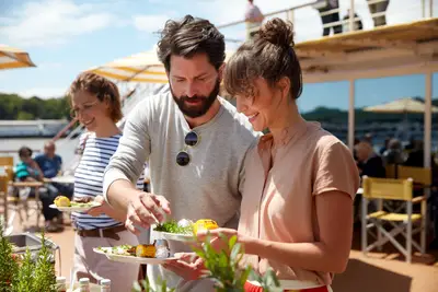 Ein Paar lächelt, während sie sich an einem Buffet auf dem Sonnendeck eines Flusskreuzfahrtschiffs bedienen. Teller mit Grillgut und Gemüse sind sichtbar.