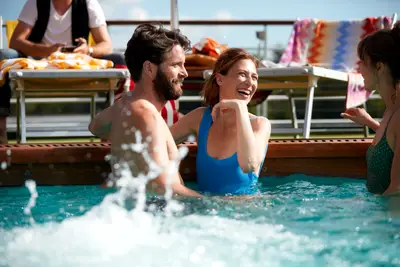Fröhliche Gruppe lacht und planscht im Pool auf dem sonnigen Sonnendeck eines A-ROSA Flusskreuzfahrtschiffs. Entspannte Urlaubsstimmung.