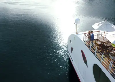 Ein elegant gekleidetes Paar steht auf dem Sonnendeck eines Schiffes und blickt auf das von der Sonne glitzernde Wasser. Im Hintergrund sind Sonnenschirme und Sitzgelegenheiten.