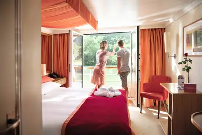 Ein Paar blickt von einem Schiffskabinenbalkon auf eine grüne Flusslandschaft. Die Kabine ist mit warmen Farben gestaltet.