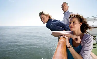 Drei Reisende, darunter zwei Frauen und ein Mann, genießen die Aussicht auf dem Sonnendeck eines Flusskreuzfahrtschiffs über das weite Meer.
