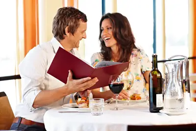 Ein lachendes Paar sitzt an einem Restauranttisch mit Wein und schön angerichtetem Essen und schaut in eine Weinkarte.