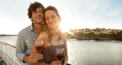 Ein lächelndes Paar steht bei Sonnenuntergang an der Reling eines Flussschiffes und hält jeweils ein Glas Rotwein. Im Hintergrund Fluss und Bäume.