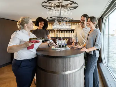 Eine A-ROSA Crew-Mitarbeiterin schenkt an der Bar Wein für lachende Gäste ein. Durchs Fenster ist der Fluss zu sehen.