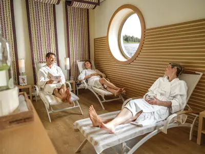 Drei Personen in Bademänteln entspannen auf Liegen im modernen SPA-Ruhebereich eines Flusskreuzfahrtschiffes mit Flussblick.