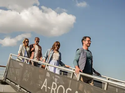 Vier Personen gehen lächelnd bei strahlendem Sonnenschein die Gangway eines A-ROSA Flussschiffs hinunter, blauer Himmel mit Wolken im Hintergrund.