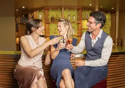 Drei Personen, zwei Frauen und ein Mann, sitzen lachend an einer schicken Bar und stoßen mit verschiedenen Cocktails an. Ein lebhafter Moment in einer Lounge.