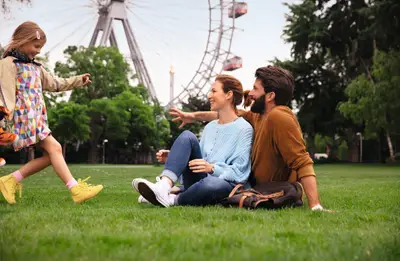Lachendes Kind spielt mit den Eltern im Wiener Praterpark auf grünem Gras, mit dem berühmten Riesenrad im Hintergrund.