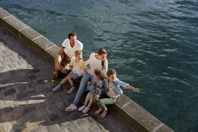 Eine glückliche Familie mit zwei Erwachsenen und drei Kindern sitzt an einem sonnigen Flussufer, ein Mädchen zeigt auf das Wasser.