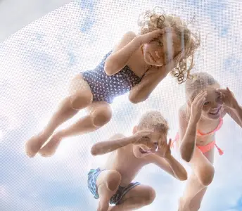 Drei fröhliche Kinder in Badekleidung schauen durch ein Glasdach, halten die Hände schützend vor die Augen. Der blaue Himmel ist durch die Perforierung sichtbar.