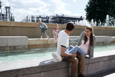 Eine Familie mit zwei Kindern spielt lachend an einem Springbrunnen im Freien, während die Eltern zusehen.