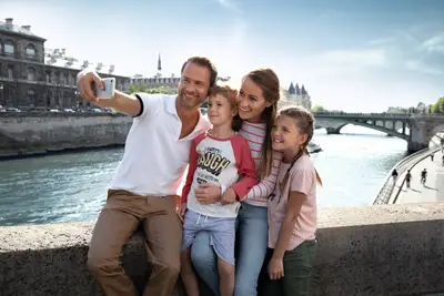 Eine glückliche vierköpfige Familie (Vater, Mutter, Sohn, Tochter) macht ein Selfie auf einer Pariser Brücke über die Seine bei sonnigem Wetter.