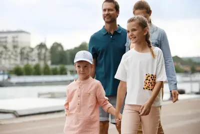 Eine lächelnde Familie mit zwei Kindern spaziert an einem Flussufer. Die Kinder halten Händchen, die Eltern folgen im Hintergrund.