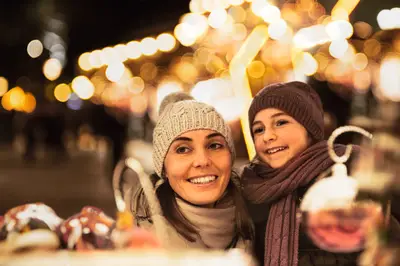 Lachende Frau und Kind in Mützen und Schals auf einem Weihnachtsmarkt bei Nacht, umgeben von funkelnden Lichtern.