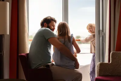 Eine Familie auf einem Flusskreuzfahrtschiff: Vater und Tochter blicken aus der Kabine auf den Balkon, wo der Sohn lachend steht.