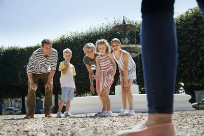 Fröhliche Familie mit drei Kindern spielt Boule auf einem sonnigen Kiesplatz vor einem Springbrunnen.