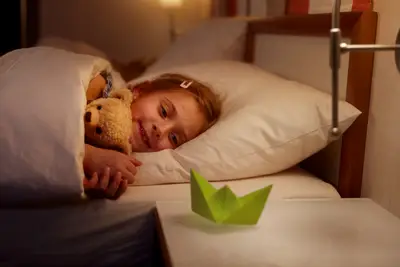 Ein lächelndes kleines Mädchen liegt mit ihrem Teddybär im Bett, warm beleuchtet, ein Origami-Boot auf dem Nachttisch.