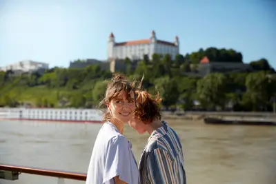 Zwei lächelnde Frauen auf einem Flusskreuzfahrtschiff blicken vom Sonnendeck auf die Burg Bratislava und die Donau.