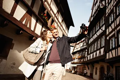 Ein glückliches Paar macht ein Selfie in einer charmanten historischen Straße mit Fachwerkhäusern in Straßburg bei Sonnenschein.