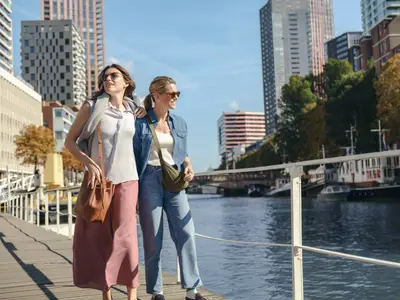 Zwei Frauen in Sonnenbrillen spazieren lächelnd an einem sonnigen Tag entlang einer Promenade mit Blick auf die moderne Skyline von Rotterdam und das Wasser.