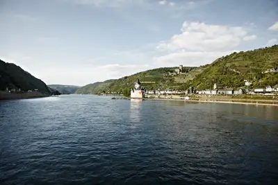 Der Rhein mit der Burg Pfalzgrafenstein und einem Ort am Ufer, umgeben von grünen Hügeln und Weinbergen unter hellem Himmel.