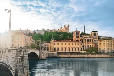 Panoramablick auf Lyon mit dem Fluss im Vordergrund, einer steinernen Brücke, historischen Gebäuden am Ufer und der Basilika Notre-Dame de Fourvière auf dem Hügel.
