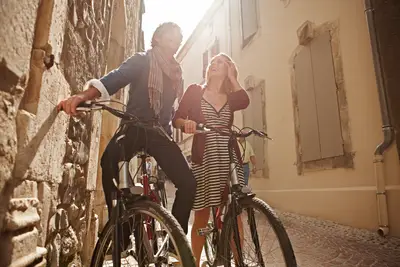 Ein glückliches Paar fährt auf Fahrrädern durch eine enge, sonnige Gasse einer Altstadt, umgeben von historischen Gebäuden.