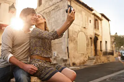 Ein lächelndes Paar macht in der Altstadt von Arles bei goldenem Sonnenlicht ein Selfie, während die Frau den Mann auf die Wange küsst.