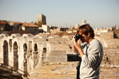 Ein Mann fotografiert bei Tageslicht die historische Stadt Arles von einer Steinmauer aus, mit alten Gebäuden im Hintergrund.