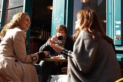 Drei Personen genießen Kaffee und Unterhaltung an einem sonnigen Café-Tisch im Freien vor einer blauen Fassade.