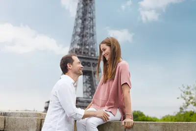 Ein lächelndes Paar in Paris, der Mann in Weiß blickt zu seiner Partnerin in Rosa auf, die auf einer Mauer sitzt. Im Hintergrund der Eiffelturm.