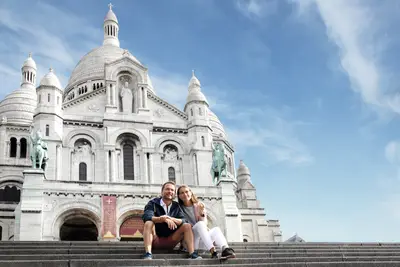Junges Paar sitzt lächelnd auf den Stufen vor der strahlend weißen Basilika Sacré-Cœur in Paris mit blauem Himmel.