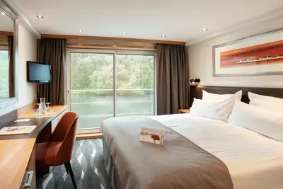 Eine komfortable Aussenkabine eines Flussschiffs mit Doppelbett, Schreibtisch und einem grossen Fenster mit Blick auf den Fluss und bewaldete Ufer.
