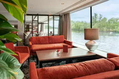 Gemütliche Panorama Lounge auf der A-ROSA Alea mit roten Sofas, einem Couchtisch und großen Fenstern mit Blick auf einen Fluss und bewaldetes Ufer.