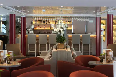 Moderner Bar-Loungebereich auf der A-ROSA ALVA mit Barhockern, beleuchteten Spirituosenregalen und roten Sesseln, dekoriert mit weißen Orchideen.
