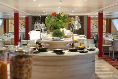 Frühstücksbuffet auf der A-ROSA ALVA mit Schalen, Tellern, Pflanzen und warmen Speisen unter Wärmelampen auf einem Holztisch.