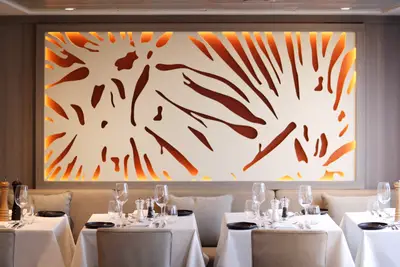 Ein elegantes Restaurant auf der A-ROSA ALVA mit weiß gedeckten Tischen, bereit für Gäste, und einer großen Wandinstallation mit abstrakten Mustern und warmer oranger Beleuchtung.