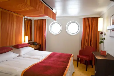 Blick in eine A-ROSA Aqua Aussenkabine mit Doppelbett, roten Kissen und Decke, orangefarbenen Vorhängen und zwei Bullaugenfenstern.