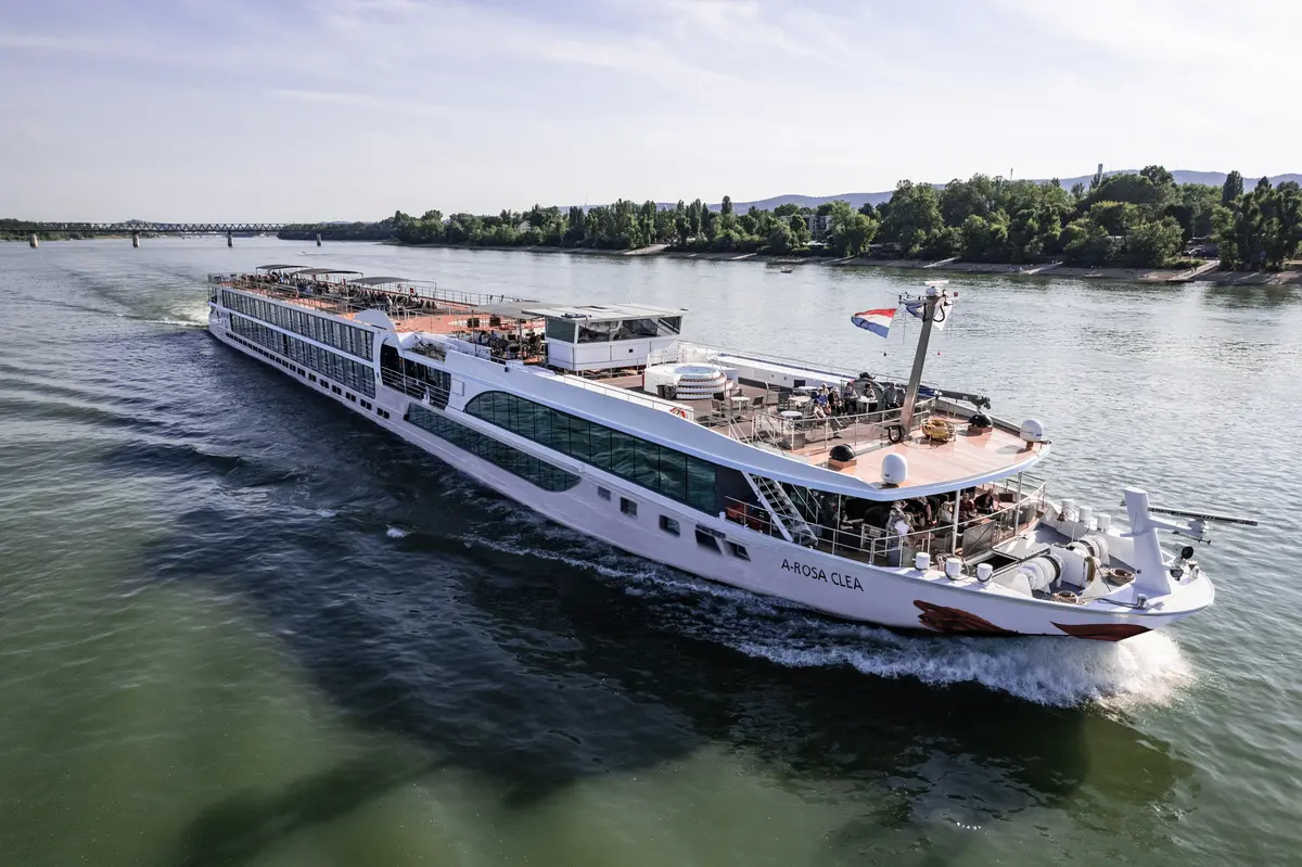 Luftaufnahme des Flussschiffs A-ROSA CLEA, das auf einem Fluss entlang eines bewaldeten Ufers fährt. Das Sonnendeck mit Whirlpool ist belebt.