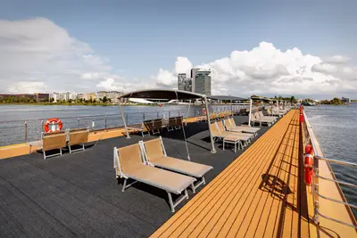 Sonnendeck der A-ROSA CLEA mit vielen Liegestühlen und Sonnensegeln unter blauem Himmel mit Wolken. Im Hintergrund die Skyline von Amsterdam.