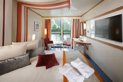Helle A-ROSA DONNA Kabine mit Doppelbett, Sesseln, Schreibtisch und Flachbildfernseher. Durchs Fenster blickt man auf Fluss und grüne Ufer. Bademäntel und Koffer sind zu sehen.