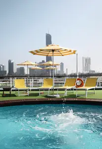 Auf dem Sonnendeck der A-ROSA DONNA sind ein blauer Pool mit spritzendem Wasser, gelbe Liegestühle und Sonnenschirme zu sehen. Im Hintergrund eine Stadtskyline.