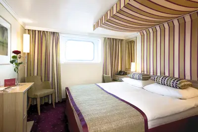 Blick in eine freundliche A-ROSA FLORA Aussenkabine mit Doppelbett, grün-lila gestreiften Vorhängen und Kopfteil, einem kleinen Fenster und einem Schreibtisch mit roter Rose.
