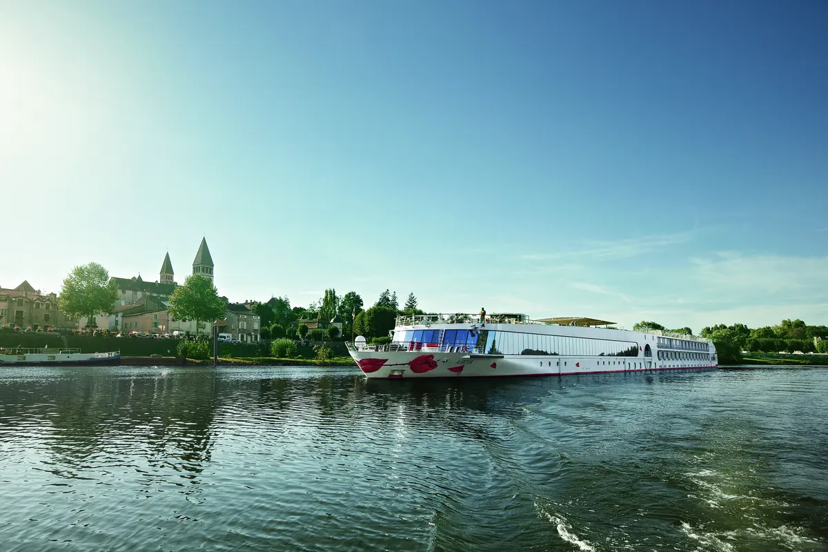 Die A-ROSA LUNA mit roter Lippengrafik am Bug fährt auf einem Fluss. Im Hintergrund ist eine historische Stadt mit einer Kirche und zwei Türmen zu sehen.