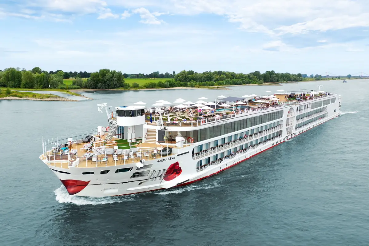 Die A-ROSA SENA, ein weißes Flussschiff mit rotem Kussmund am Bug, fährt auf dem Rhein. Das Sonnendeck ist mit vielen Gästen gefüllt.