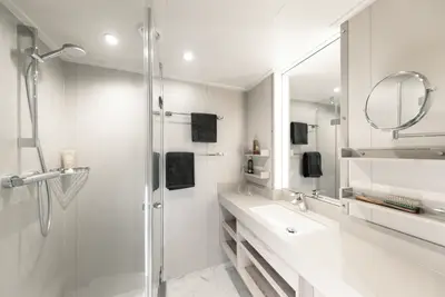 Modernes Badezimmer einer A-ROSA SENA Schiffskabine mit begehbarer Dusche, Waschtisch, beleuchtetem Spiegel und dunklen Handtüchern.