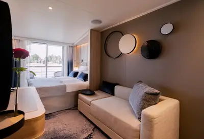 Innenansicht einer modernen A-ROSA SENA Balkonkabine mit hellem Sofa, Doppelbett, Design-Wanddeko und Flussblick.