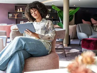 Eine Frau mit Locken sitzt entspannt in einer modernen Schiffs-Lounge und bedient ein Tablet. Im Hintergrund sind weitere Sitzgelegenheiten und eine große Pflanze zu sehen.